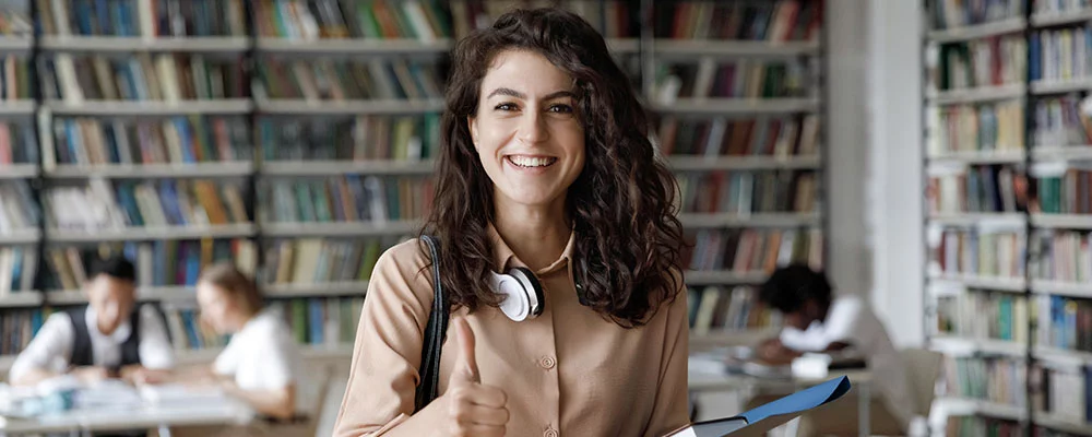 Attraktive Studentin 18s Mädchen in der Bibliothek stehen Daumen hoch, genießen interessante effektive Studien in höheren Institutionen. Erfolgreicher exzellenter Lerner, Stipendium, qualitativ hochwertiges Bildungskonzept