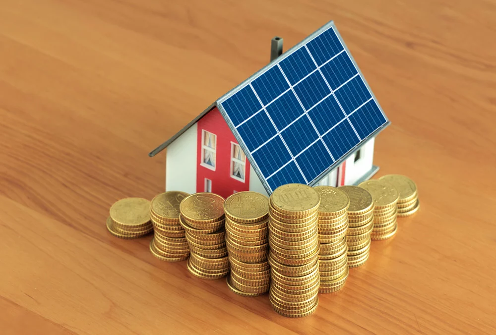 Abbildung eines kleinen Modellhauses mit Solarzellen auf dem Dach und mehrere Stapel Geldmünzen davor. Symbolbild für teure energetische Gebäudesanierung.