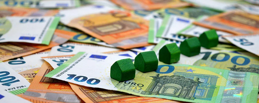 50 und 100 Euro Scheine liegen verteilt auf einem Tisch, auf dem Geld stehen kleine Bauklötze in Form von Häusern